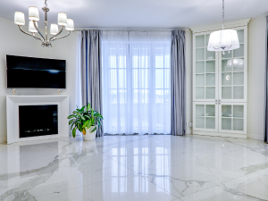 Bright modern living room with a wall-mounted TV above a white fireplace, a chandelier, and a large window with sheer curtains. A potted plant sits beside the fireplace.